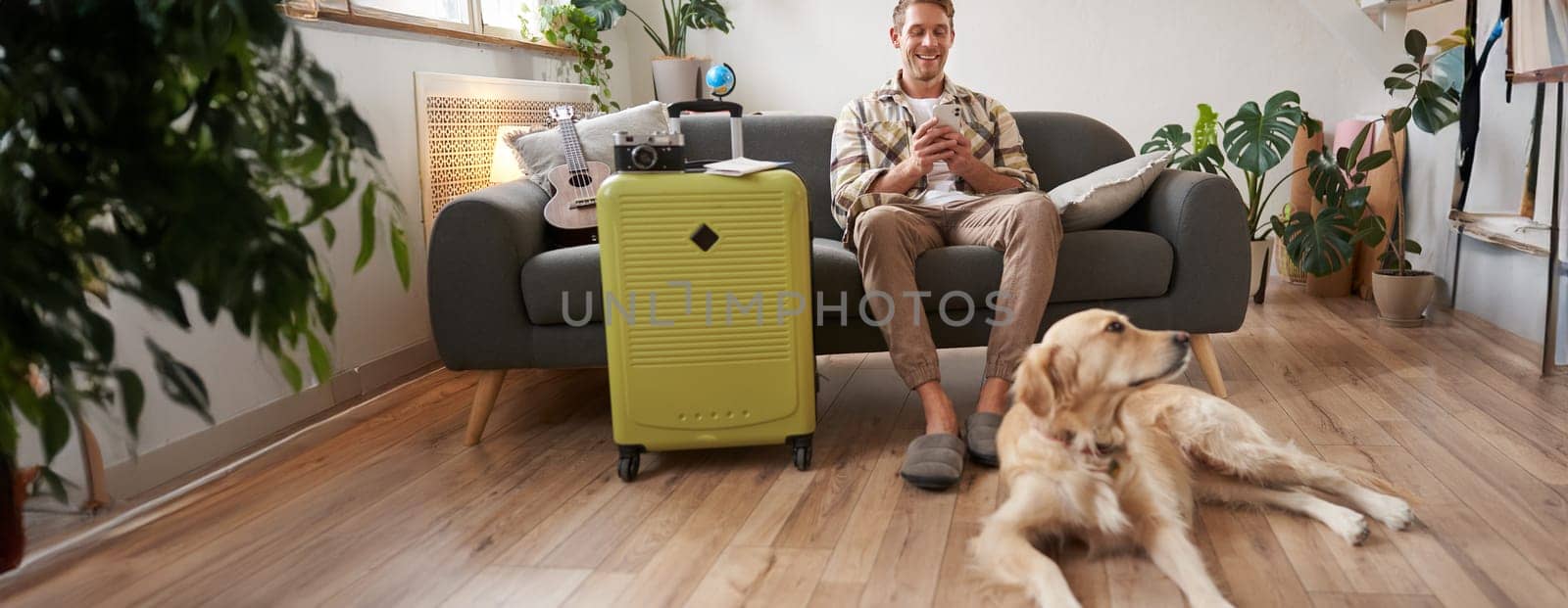 Portrait of happy, handsome young man with suitcase, sitting with his dog in living room, prepared to go on vacation, going on holiday. Tourism concept