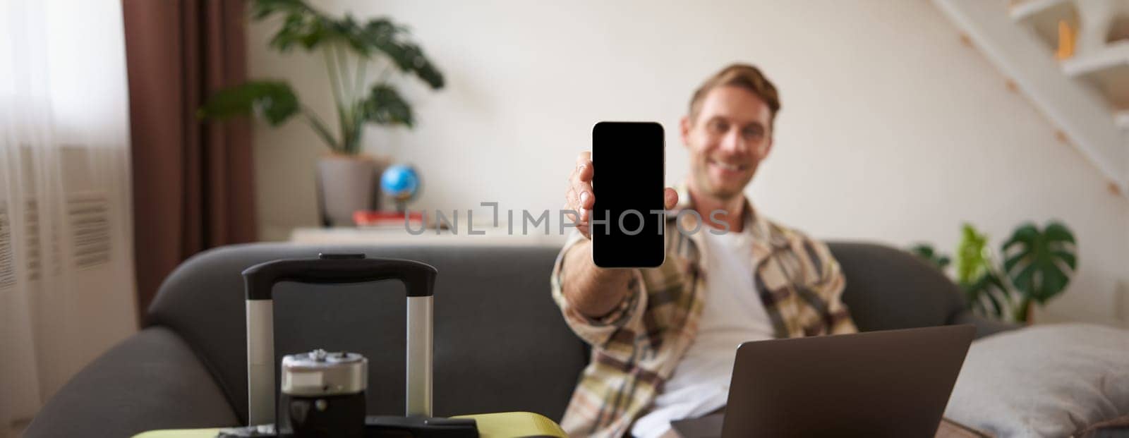 Portrait of handsome young man, showing smartphone screen, recommending mobile app for travelling or booking online, sitting with laptop and suitcase. Copy space