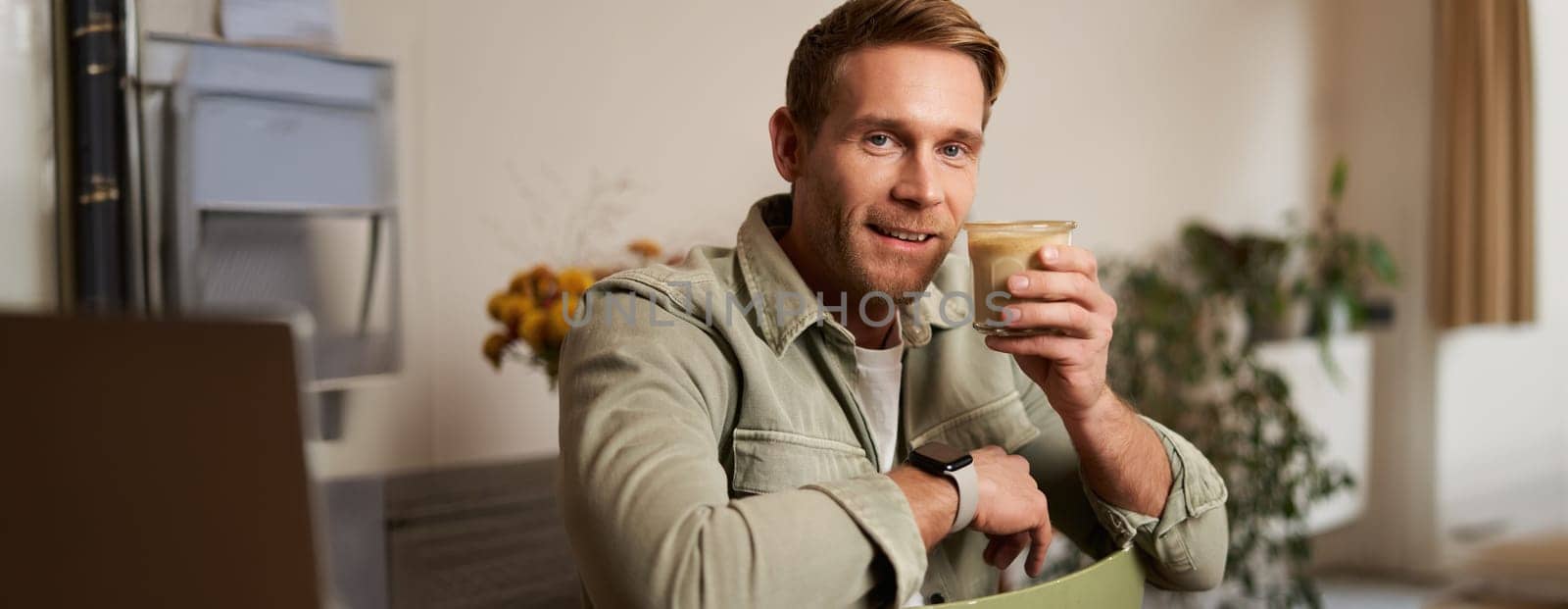 Portrait of good-looking young man with cup of coffee, sitting on chair in cafe, smiling and relaxing with his cappuccino drink.