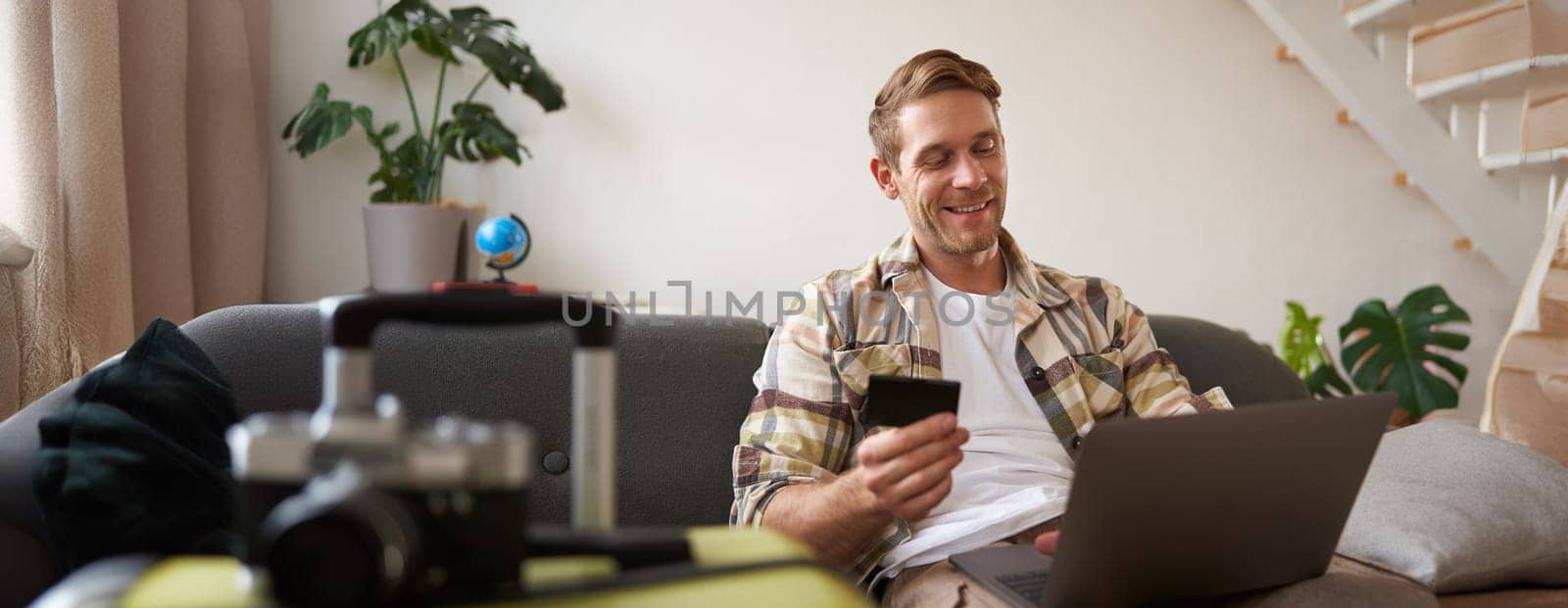 Smiling handsome guy with credit card and laptop, tourist booking a hotel for holiday, sits near suitcase.
