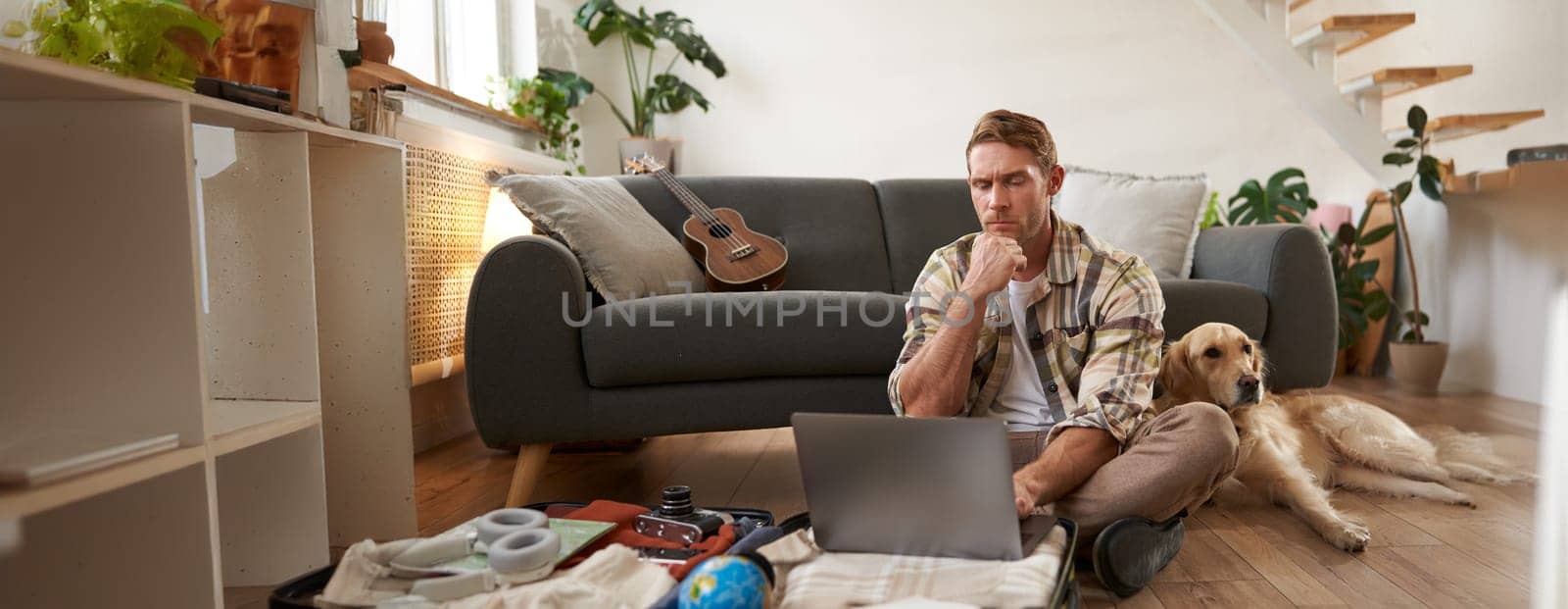 Image of young man, tourist going on holiday, sitting with a dog in living room, looking at laptop, check-in on a flight on computer. Tourism and travelling concept
