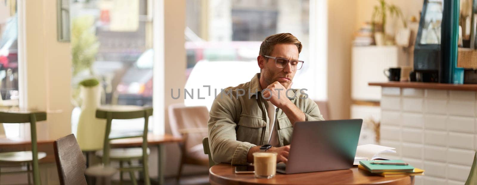 Portrait of businessman in glasses, sits in cafe with laptop, looks concentrated at screen with his project task, works remotely from coffee shop.
