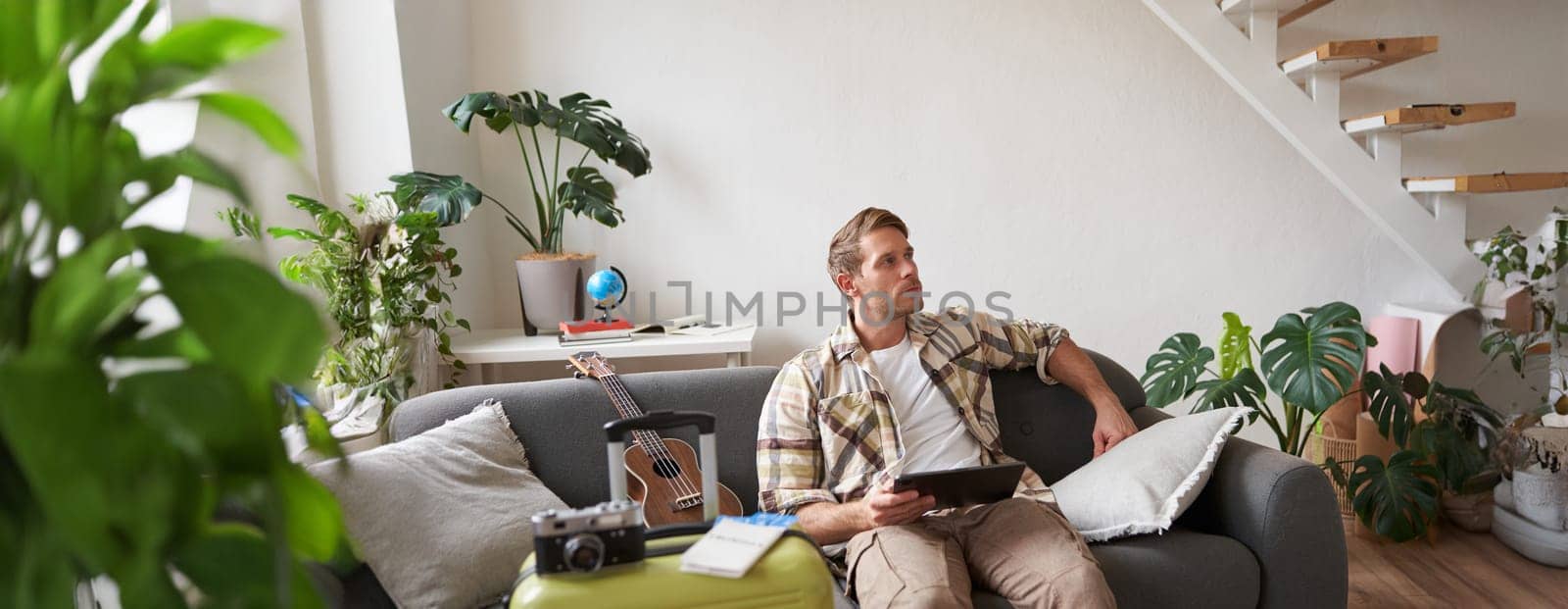 Portrait of handsome young man, tourist sitting with digital tablet near suitcase and two tickets, booking something online for his vacation, preparing for holiday.