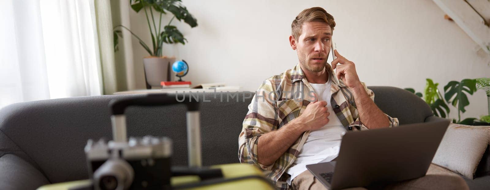 Man with confused face, talks on mobile phone, sits with laptop, has packed suitcase near him, enquires about holiday with travel agency.