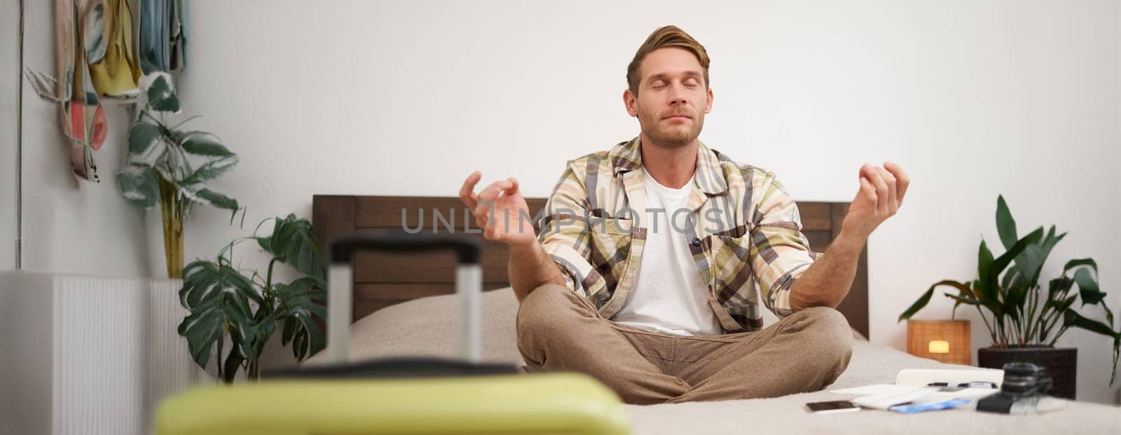 Close up portrait of meditating tourist, man sits with suitcase in bedroom, practice yoga, relaxing, finding nirvana. Tourism and travelling concept