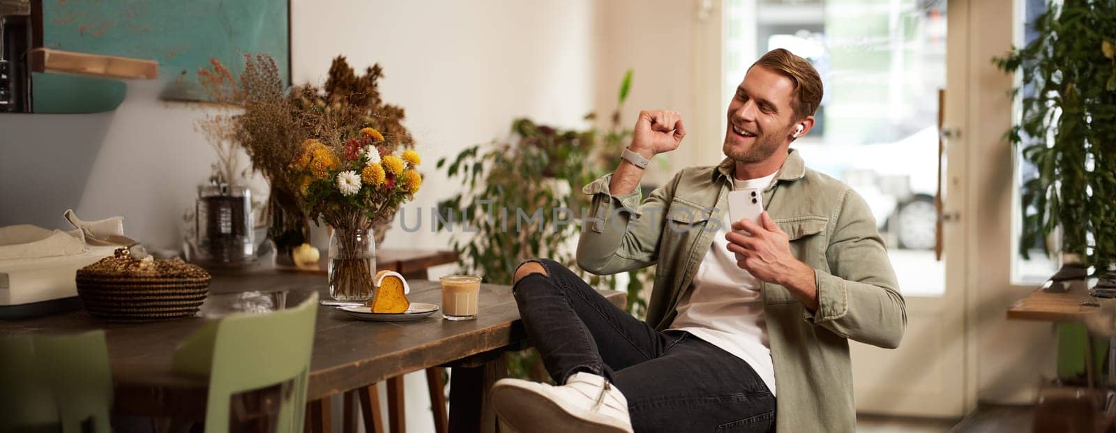 Portrait of handsome happy man in cafe, listens to music in wireless earphones, holding smartphone, connects to public wifi and enjoys favourite song, relaxing in coffee shop.