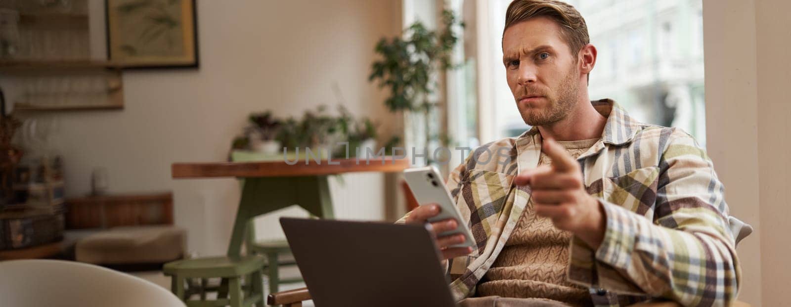 Portrait of man with serious face, sitting in cafe with laptop and smartphone, pointing at you with warning, scolding you, frowning with disapproval.