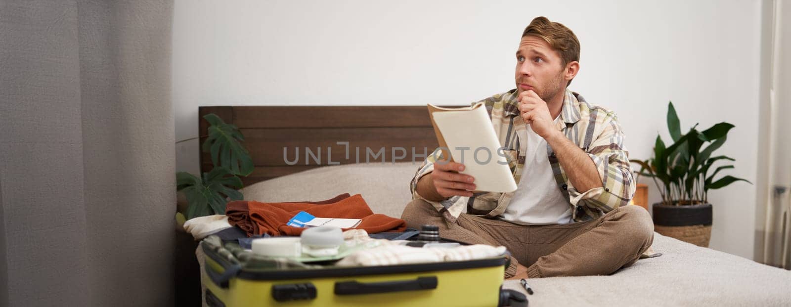 Portrait of thoughtful young man, tourist planning his vacation, sitting with open suitcase on bed, making notes, writing to do check list in notebook.