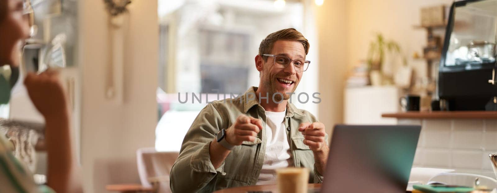 Portrait of cheerful guy in glasses, sits in cafe and works on laptop, pointing fingers at camera, inviting you, recommends you something, promoting product or public space.