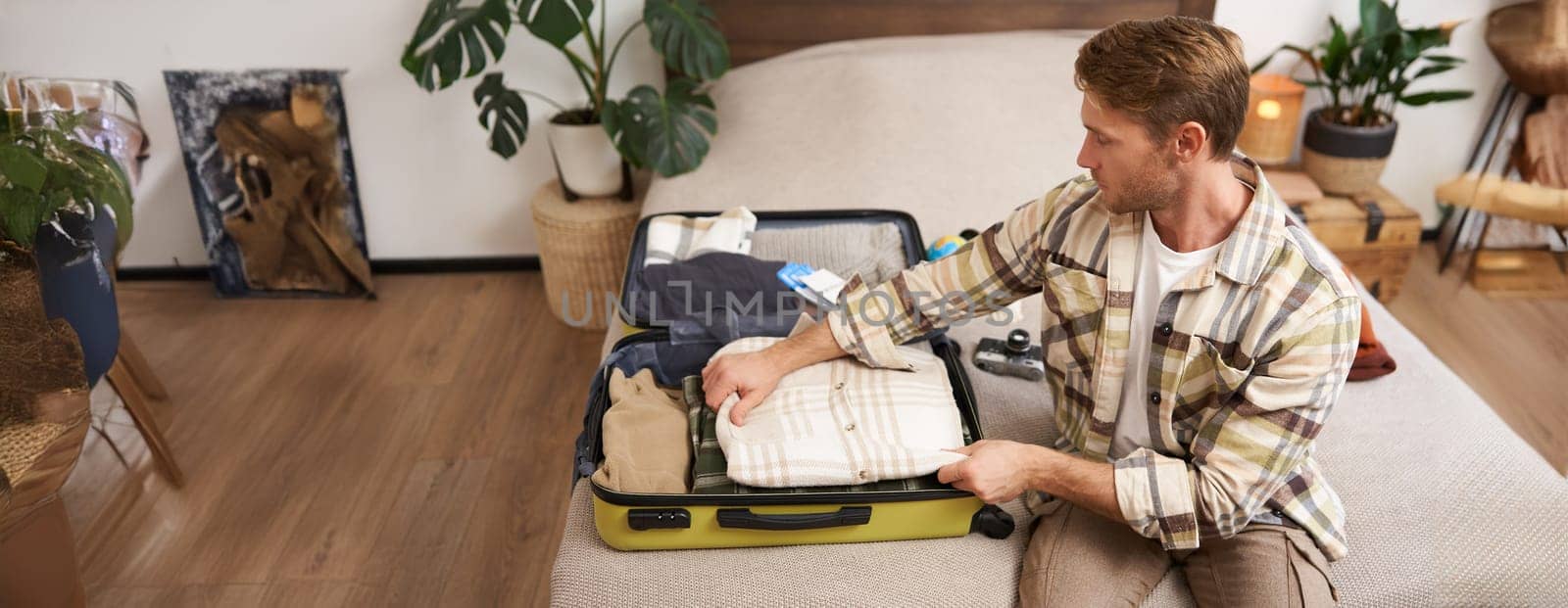 Tourism and travelling concept. Portrait of handsome young man going on holiday, packing his suitcase, checking luggage, sitting on bed and looking at packed things.