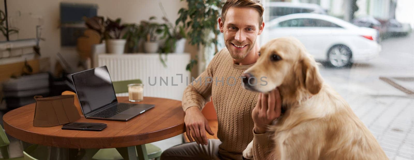 Close up portrait of smiling handsome european man with his dog in a cafe. Guy pets his golden retriever while working outdoors in coffee shop.