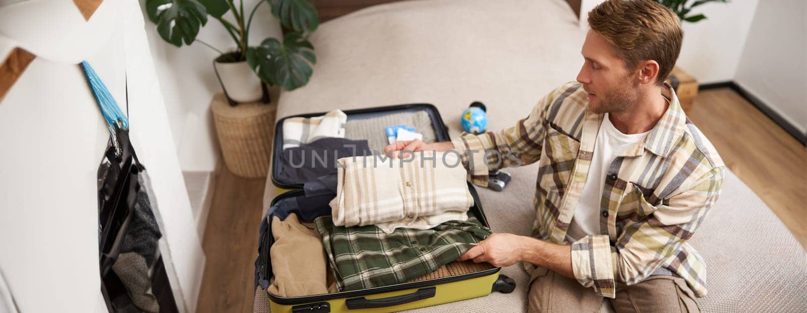 Portrait of young handsome man, tourist going on vacation, packing luggage for holidays, putting clothes in suitcase, sitting on bed.