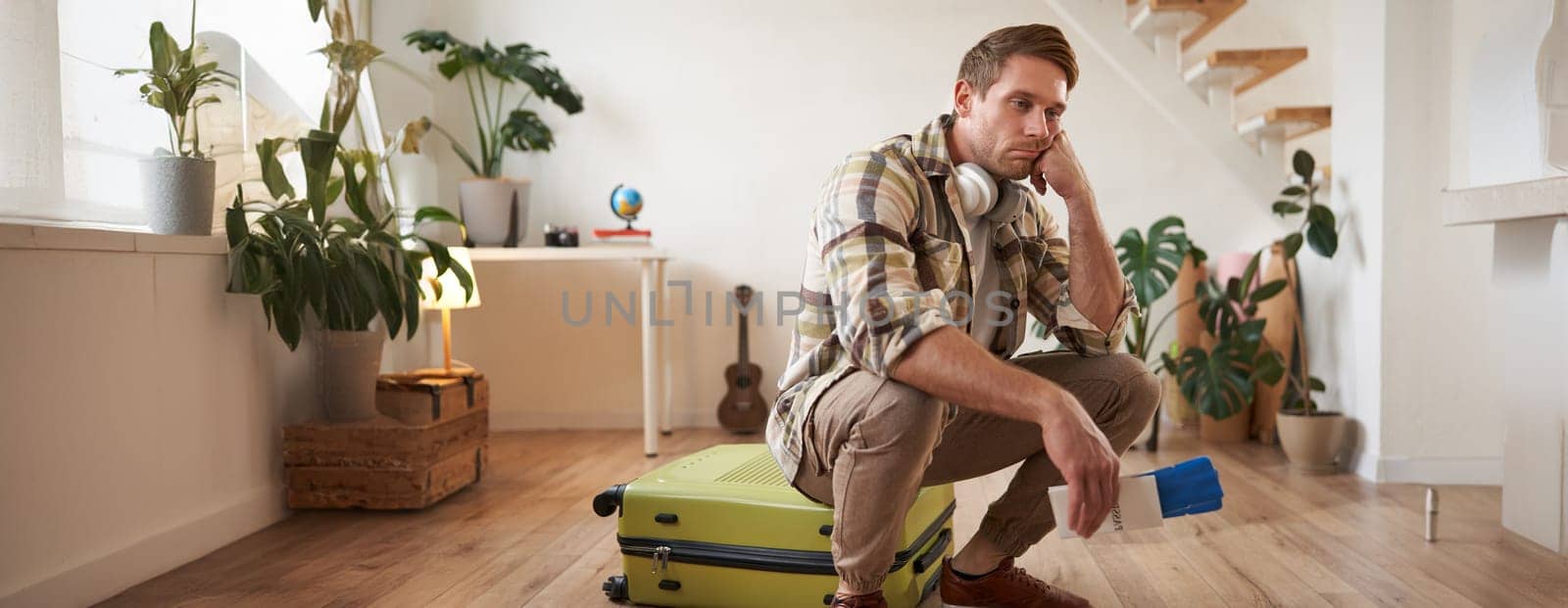 Portrait of sad and tired man, sits on his suitcase, holds passport and flight tickets, looking very upset.