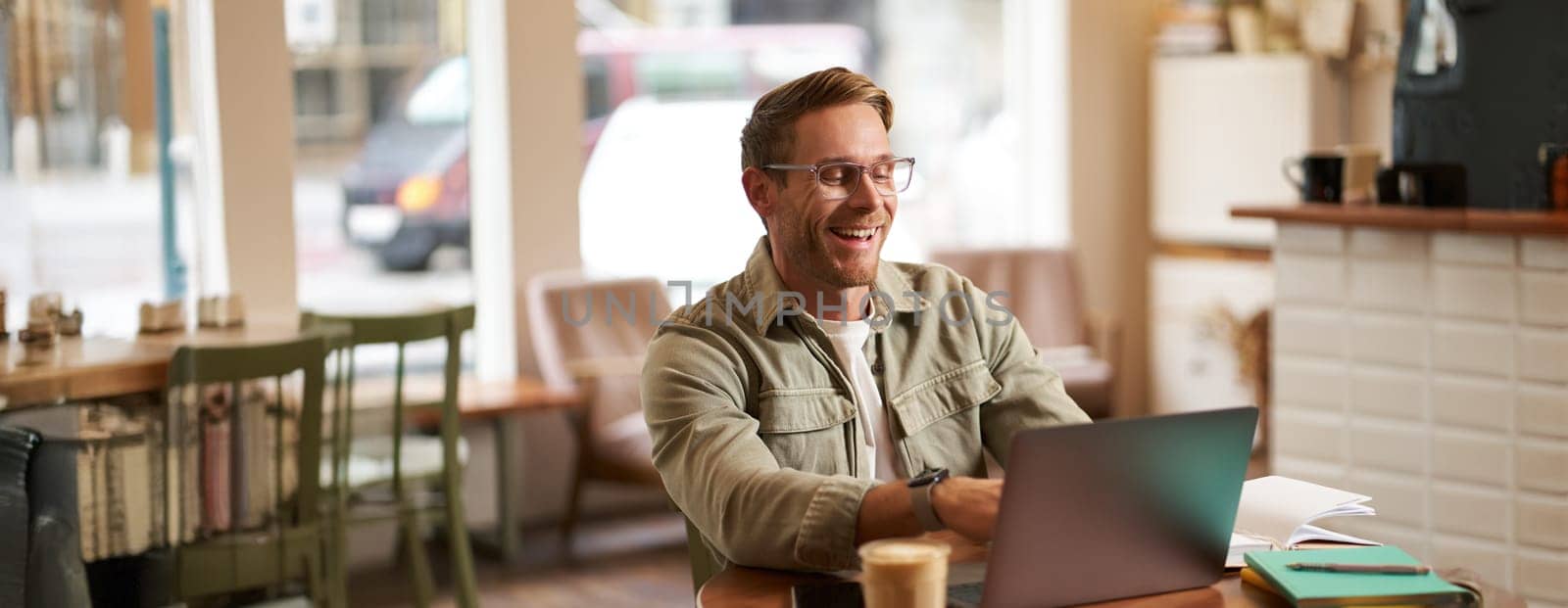 Image of young digital nomad, man in glasses sits in cafe, works from coffee shop, uses laptop in co-working space, wears glasses, drinks his beverage.