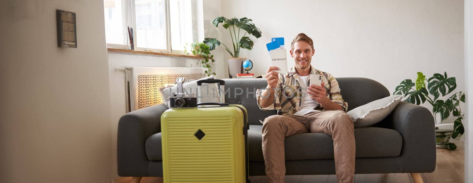 Image of handsome male model, tourist with suitcase, shows his two tickets on plane, uses mobile phone app, sits on sofa. Tourism and travelling concept