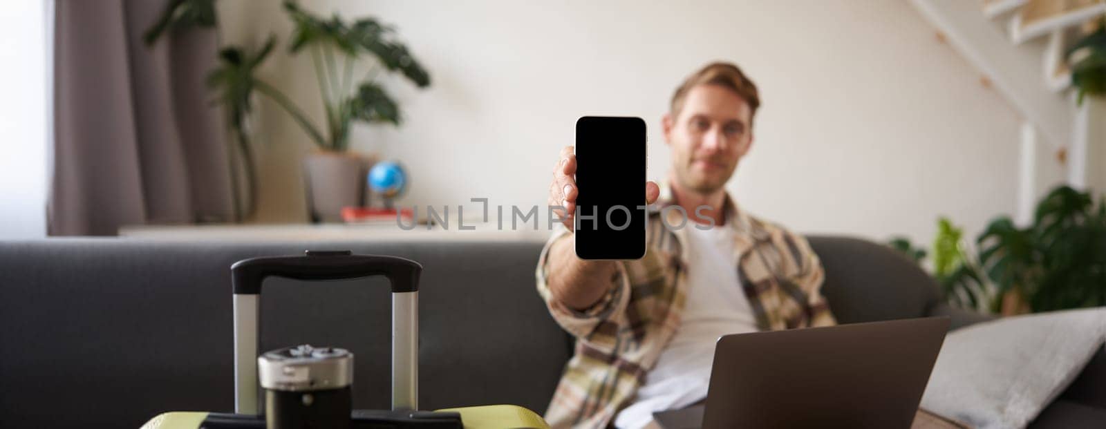 Portrait of man, tourist shows his mobile phone screen, recommends travel app, sitting with suitcase and laptop.