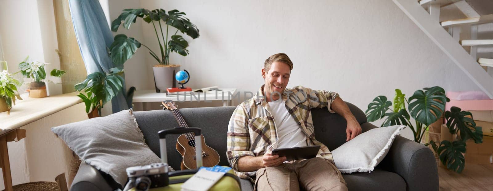 Image of young man, tourist goes on holiday, packed suitcase, sits with digital tablet on sofa, reads, looks at his map, books hotel for business trip. Copy space