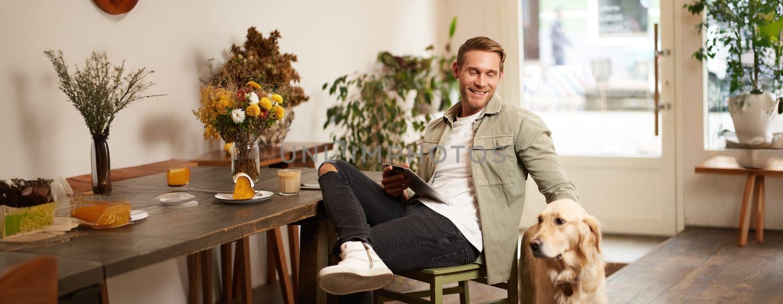 Portrait of happy smiling young man, cafe visitor, sitting in coffee shop with his dog, petting golden retriever, holding digital tablet, reading news or browsing on internet.