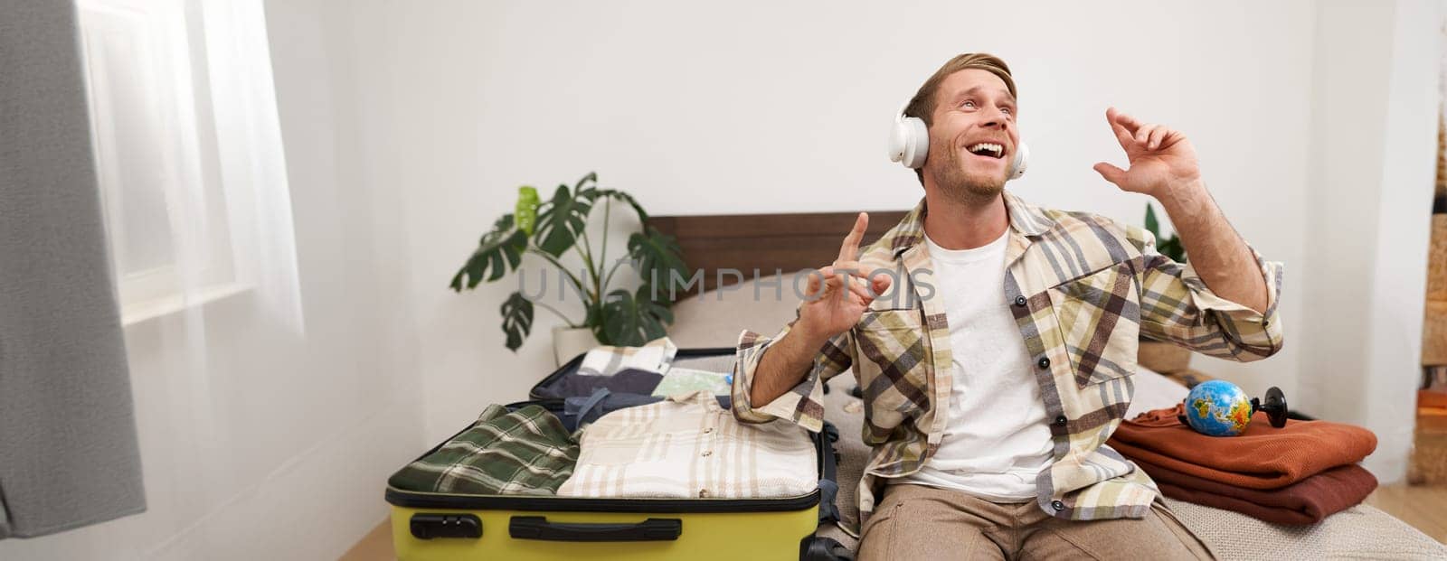 Portrait of cheerful handsome man, tourist going on vacation, dancing and listening to music in headphones while packing suitcase with clothes for holiday trip abroad.