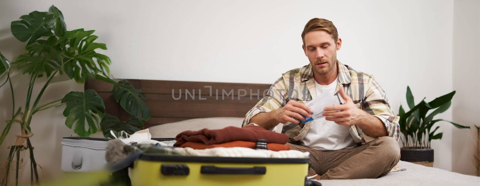 Portrait of handsome man, tourist, looking at his holiday tickets, booked vacation with travel agency, sitting on bed and packing clothes in suitcase.