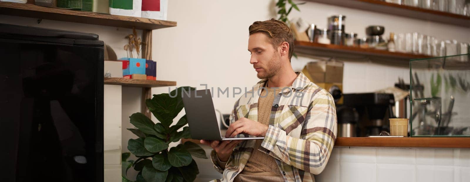 Image of young stylish guy with laptop, sitting in cafe with his computer, typing something, drinking coffee and doing project.