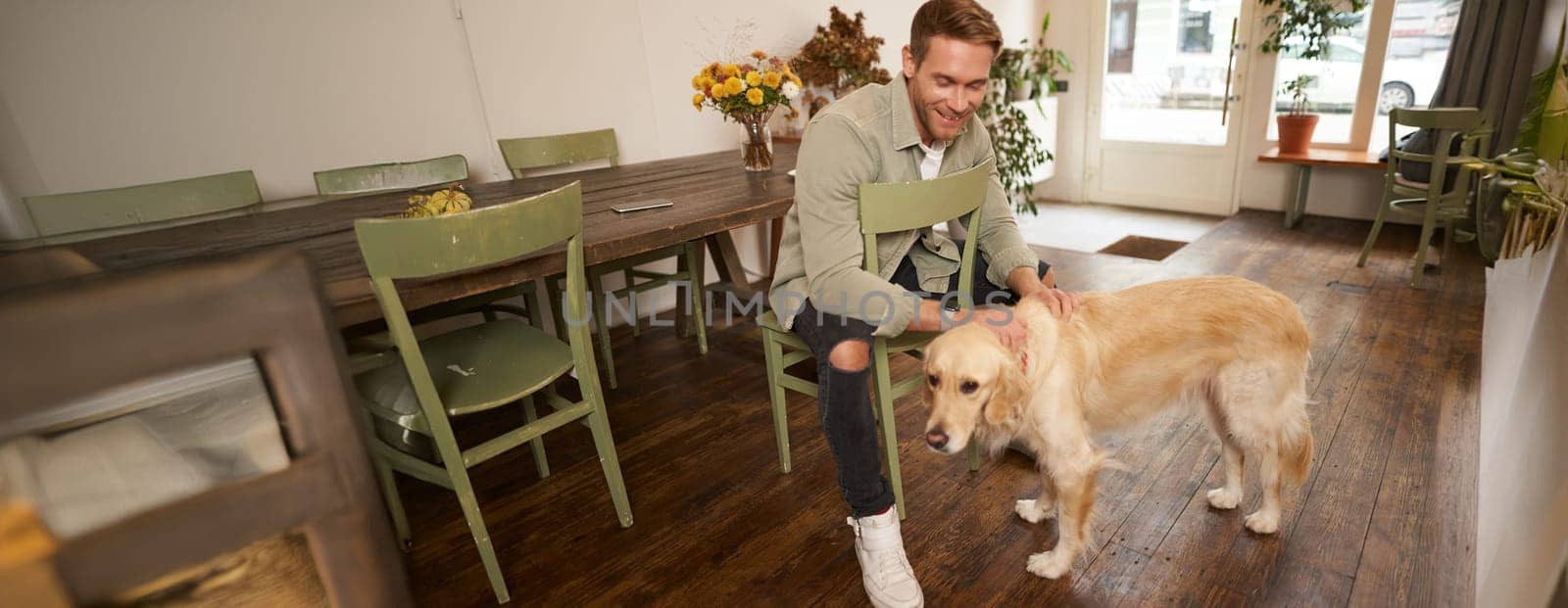 Close up portrait of handsome young man waiting for his order in coffee shop, petting a dog, resting in pet-friendly cafe.
