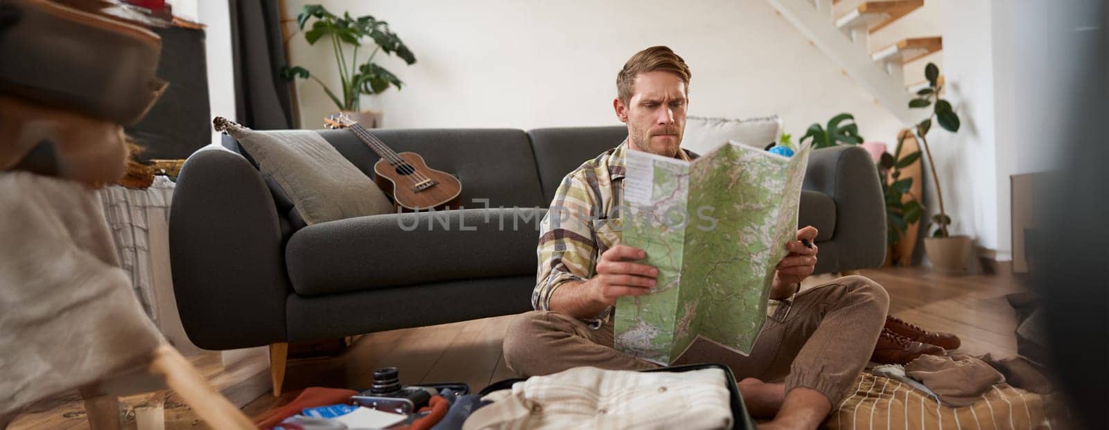 Portrait of young man, traveller, sitting packing suitcase, looking at travel map, choosing holiday destination, studying the route on vacation. Tourism concept