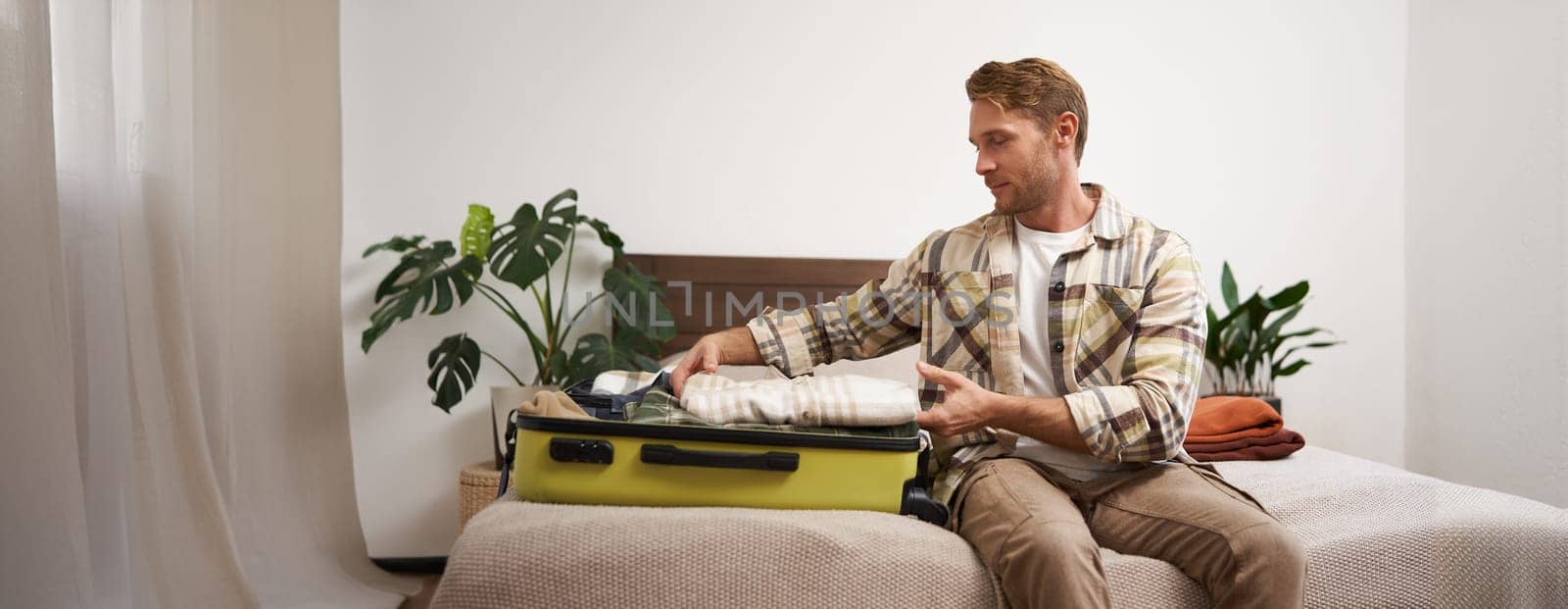 Portrait of young man packing his luggage for a trip, going on holiday, sitting on bed and putting clothes in suitcase, preparing for his flight.