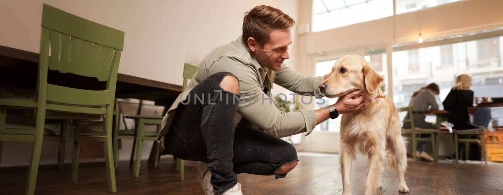 Close up portrait of handsome happy man petting his cute dog in a pet-friendly cafe. Coffee shop visitor with his golden retriever.