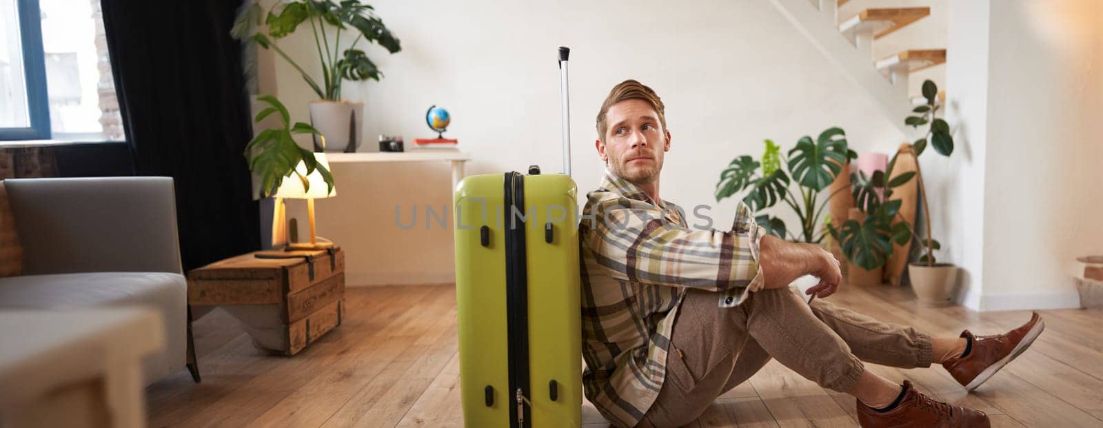 Portrait of male model with suitcase, sitting on floor, holding flight tickets and passport, going abroad, waiting for a taxi to airport.