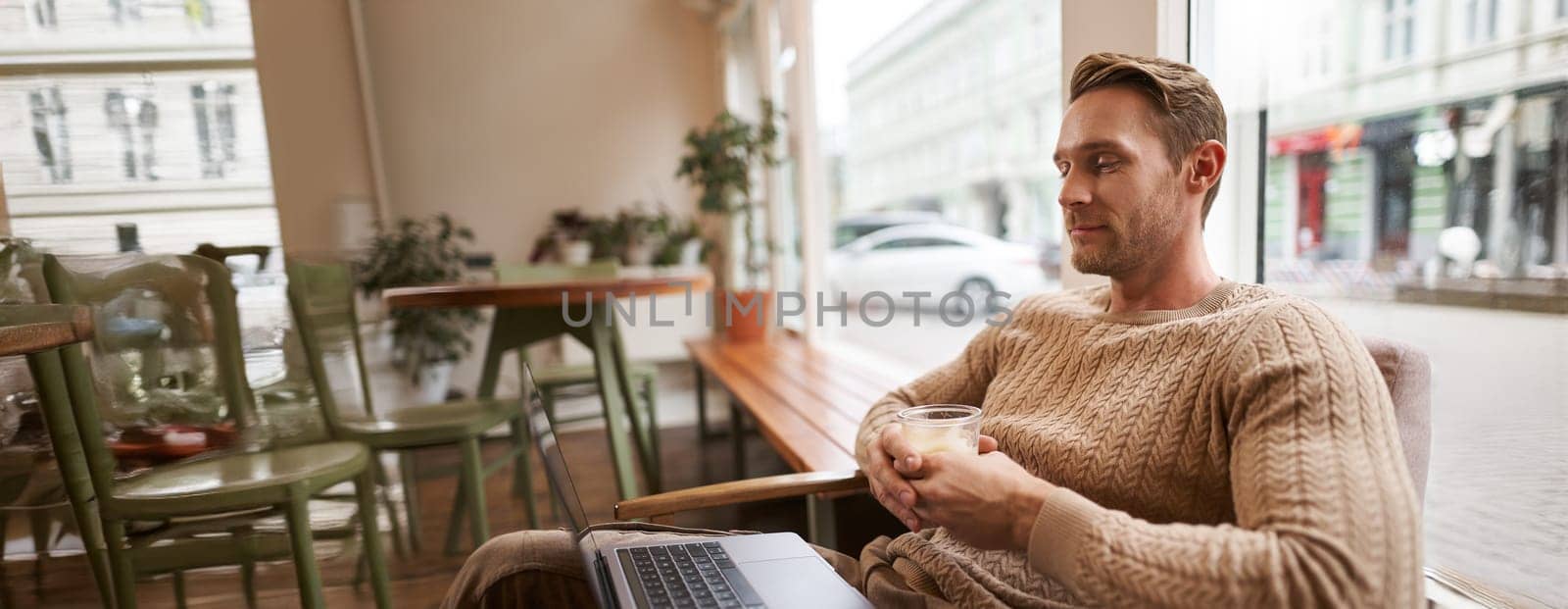 Portrait of male entrepreneur, digital nomad sitting in cafe with glass of cappuccino, looking at laptop, watching video, attending meeting online from coffee shop.