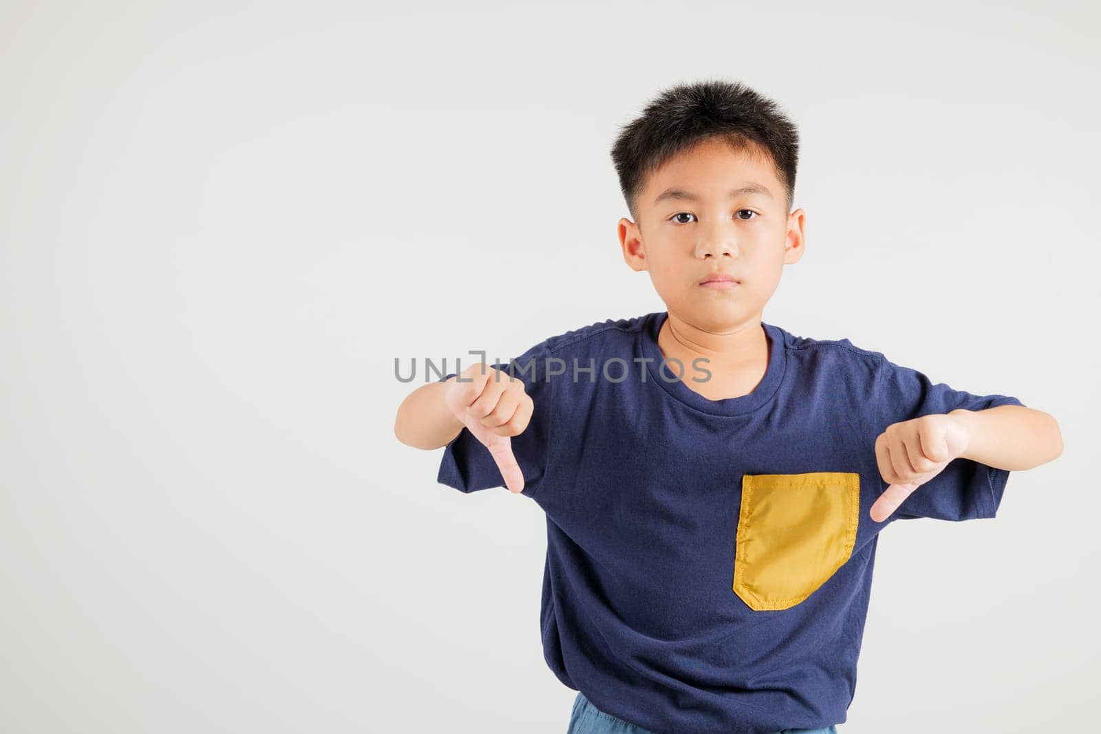 Portrait disheartened Asian young kid boy displaying a thumbs down gesture, symbolizing dislike. Studio shot isolated on white with ample copy space, primary child portraying negative and sad look