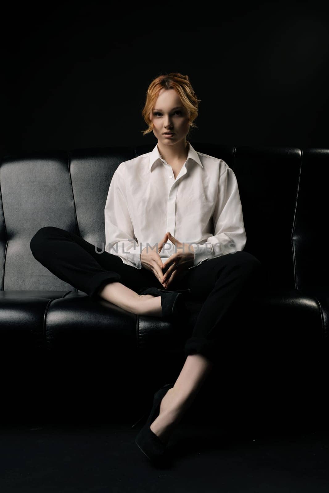 Full body of confident serious young female manager with ginger hair in elegant white shirt sitting on comfortable leather sofa - hand together against black background