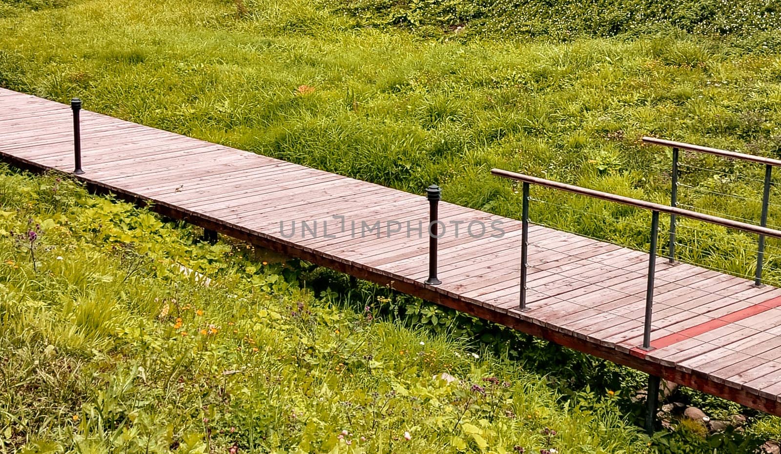 Wooden modern footpath with lanterns, along a green meadow, across a ravine, on a sunny summer day.