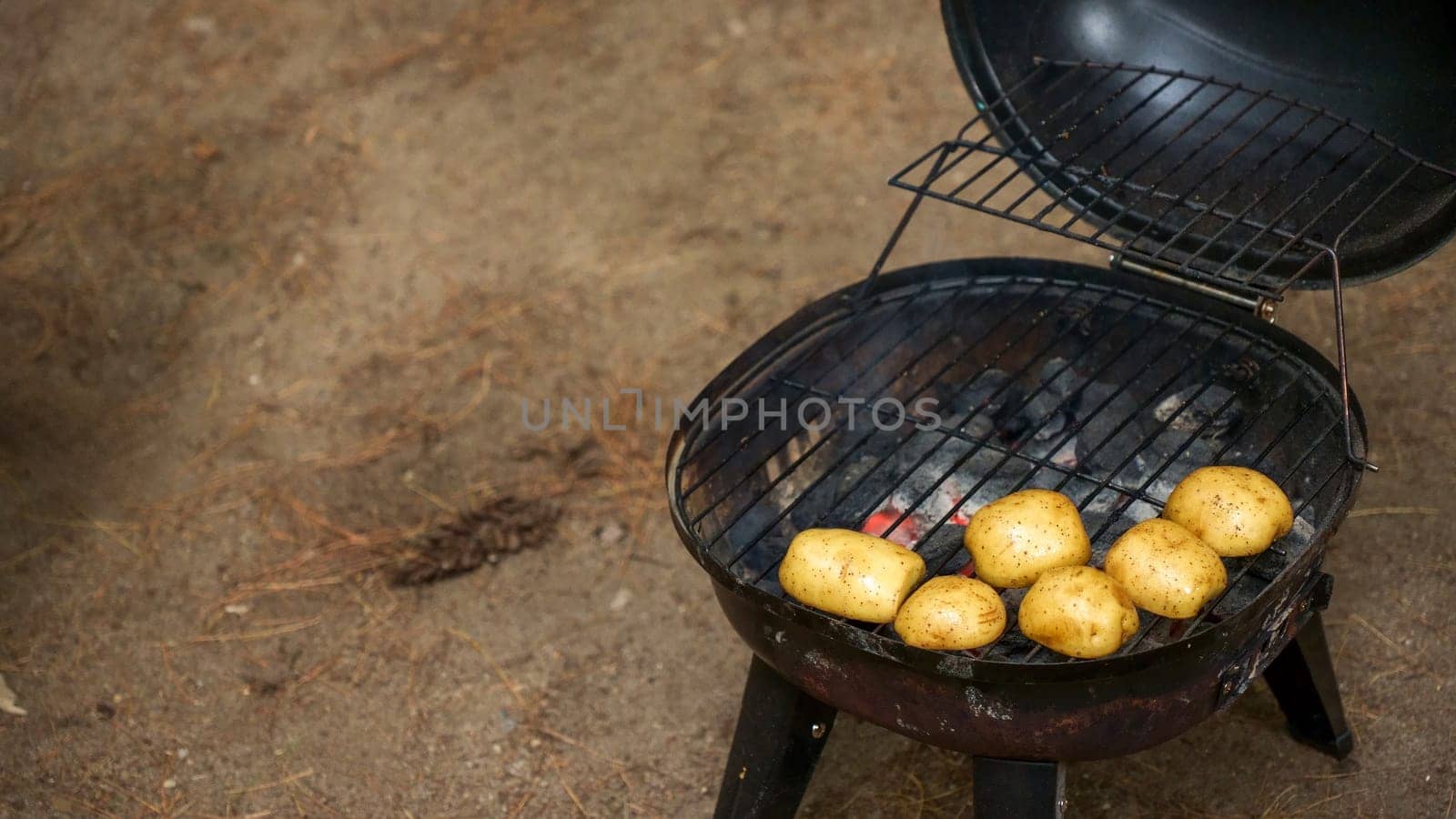 A selection of unwashed potatoes are placed on a charcoal grill, cooking with the lid open. The smoky aroma fills the air as the potatoes sizzle on the grate, showcasing a delicious grilling process.