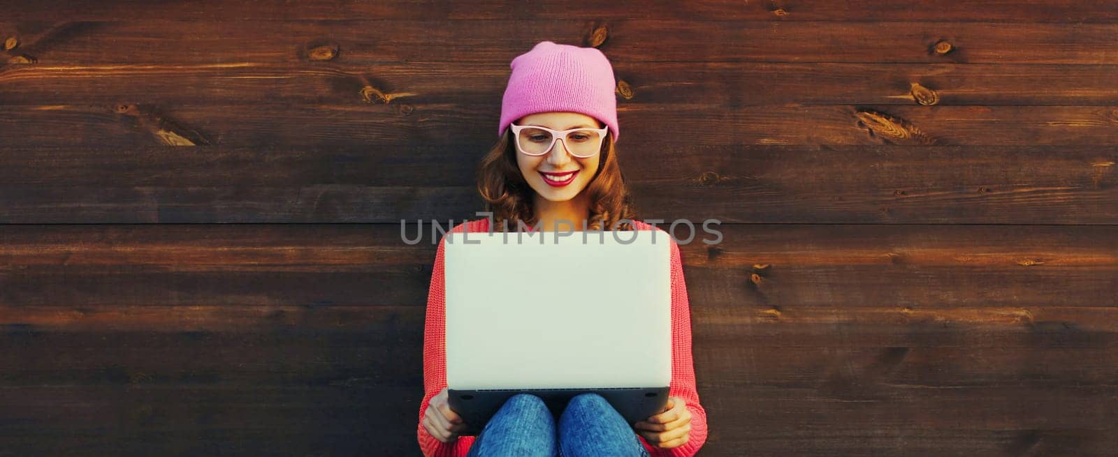 Happy modern young woman working on a laptop, freelancer girl looking at computer with glasses sitting in urban setting on wooden wall background