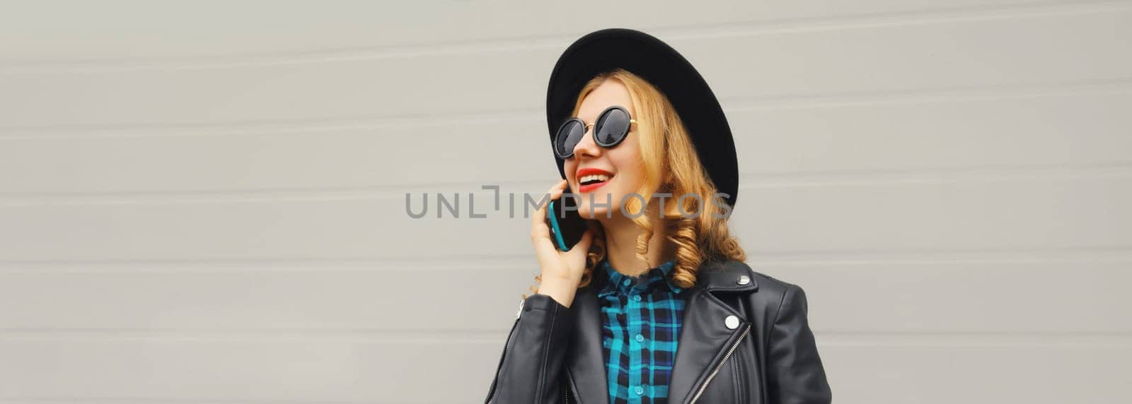Beautiful happy smiling young woman calling on phone, stylish girl talking on the smartphone in black hat, jacket on gray background
