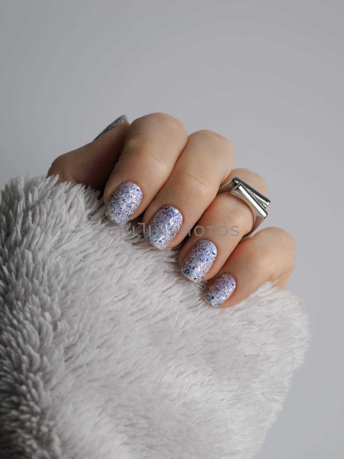 Stylish close-up of white crelly nail polish with blue shimmer, aurora flakes adorned with black foil pieces featuring silver ring and sleeve of light gray fleece sweater in neutral palette