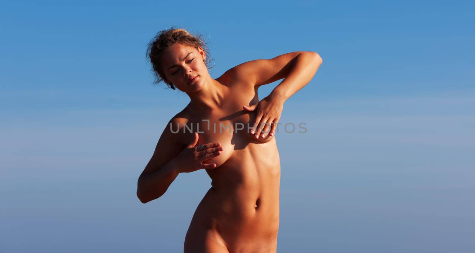 A beautiful nude woman, with fair skin, stands against a bright blue sky, shielding her breasts with hands.