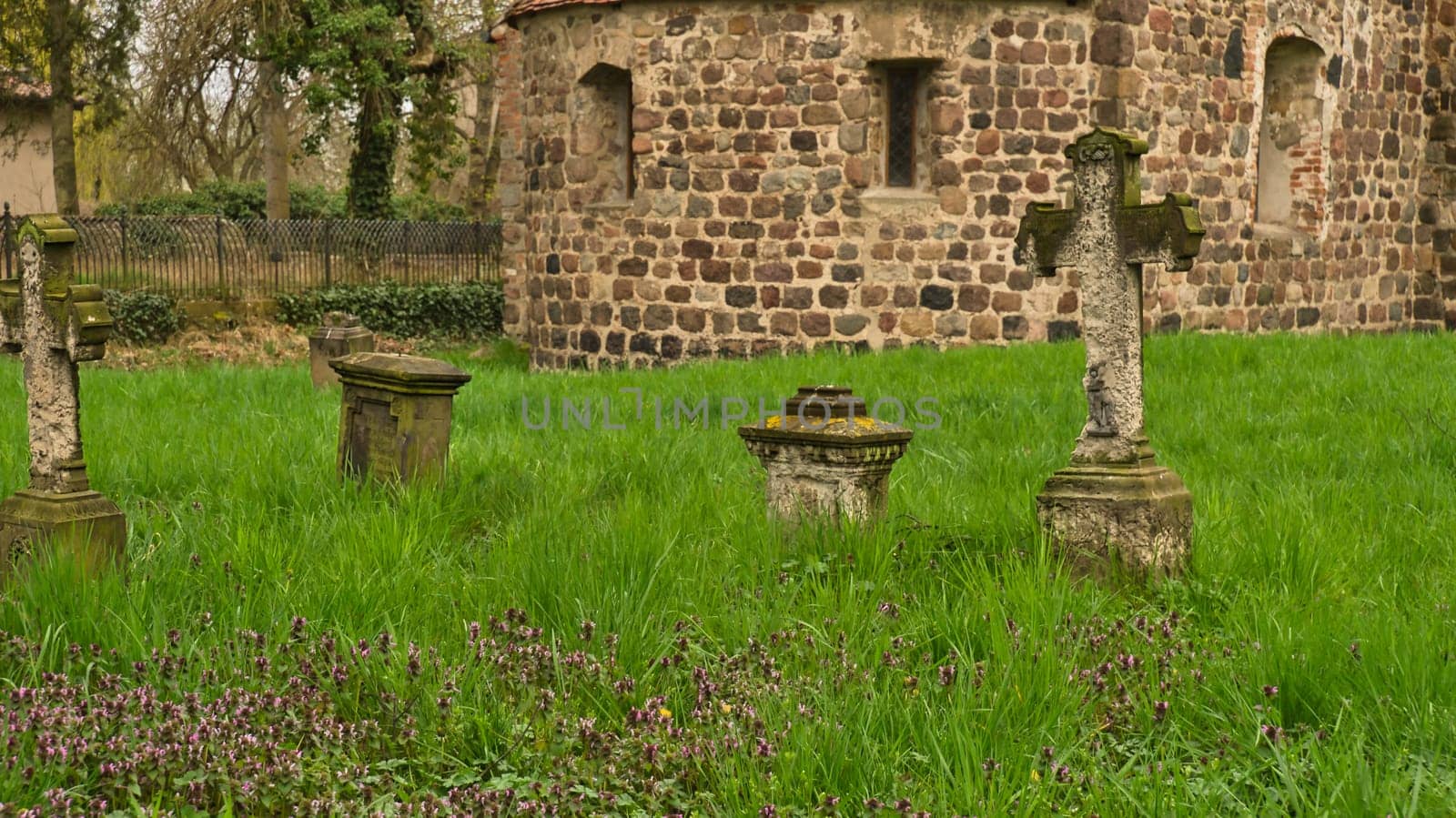 A historic stone cross in front of an old rustic church with small windows. Lively green grass in the foreground adds depth and contrast to the picture.