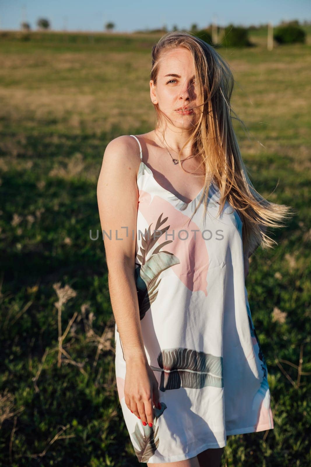 Portrait of a blonde woman in a summer dress on a green field at sunset