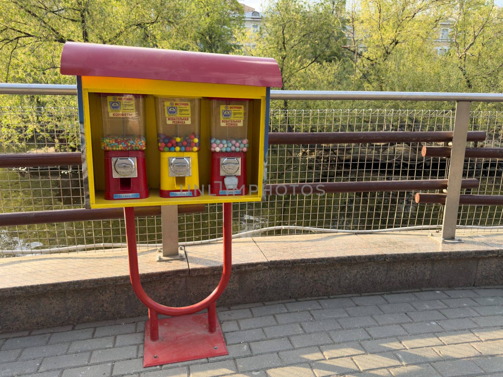 Bright vending machines with candy and chewing gum stand on city promenade by river. Cheerful colors contrast with greenery and calm water in background, evoking childhood memories
