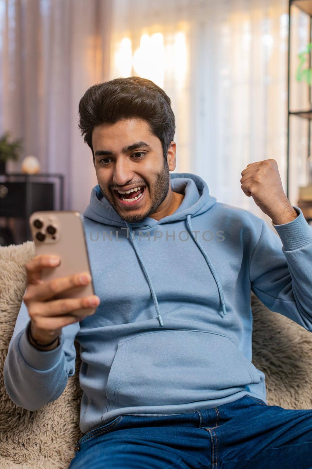 Surprised excited Indian young man holding smartphone, amazed by online game win or good news. Happy guy on sofa overjoyed by success, raises arms triumphantly, celebrating victory in living room.