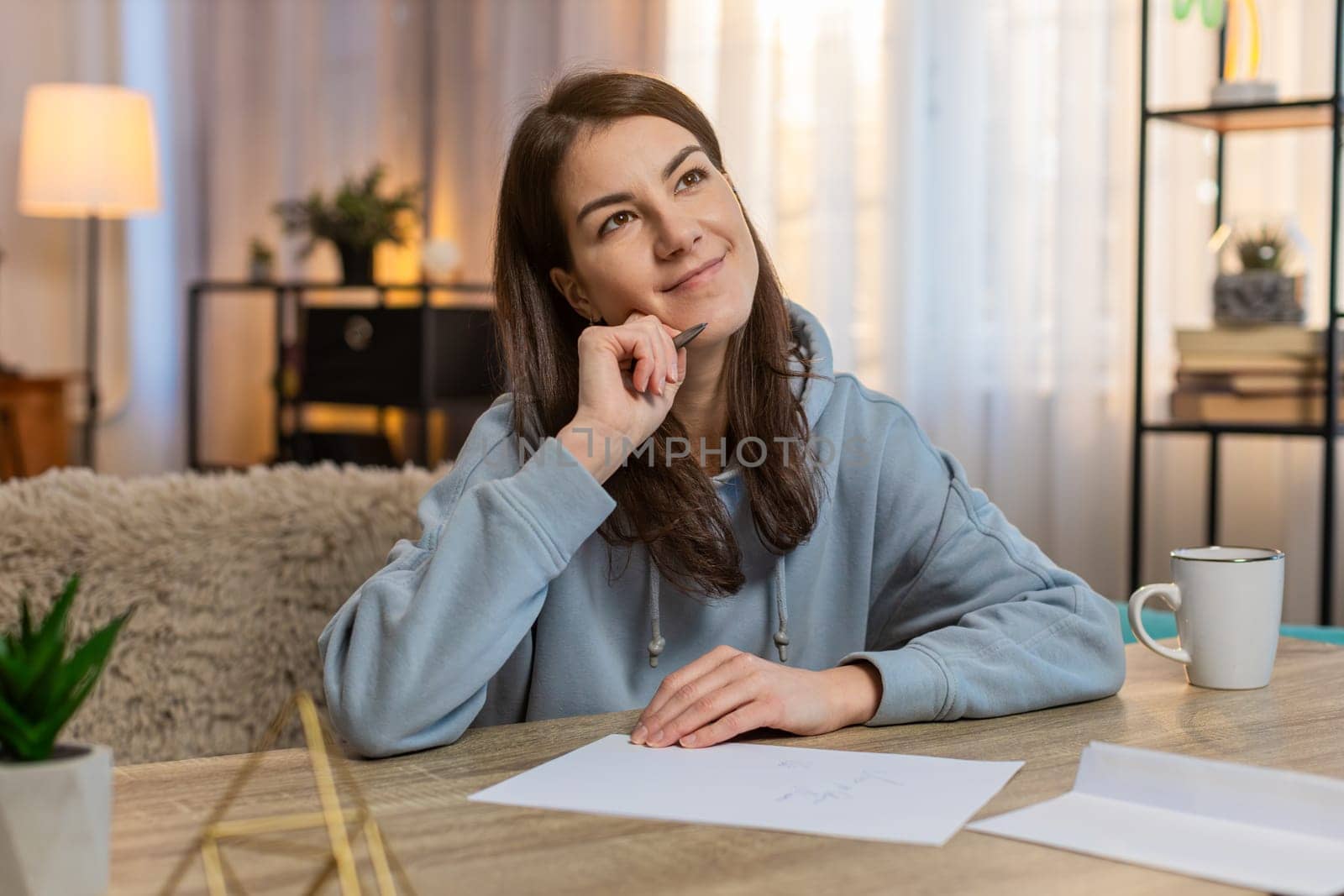 Attractive young Caucasian woman sitting on comfortable sofa at table deeply focused on writing important letter by hand. Envelope lies nearby, suggesting she is preparing it for sending or delivery.