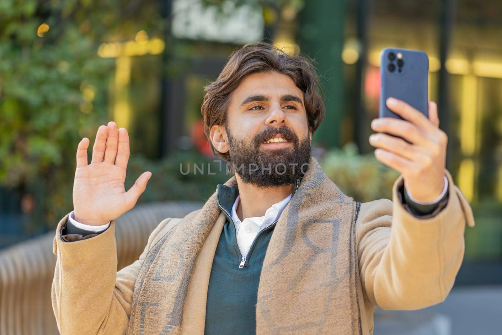 Indian young businessman taking selfie on smartphone, communicating video call online with subscribers, recording stories for social media vlog. Lebanese bearded guy blogger on downtown city street