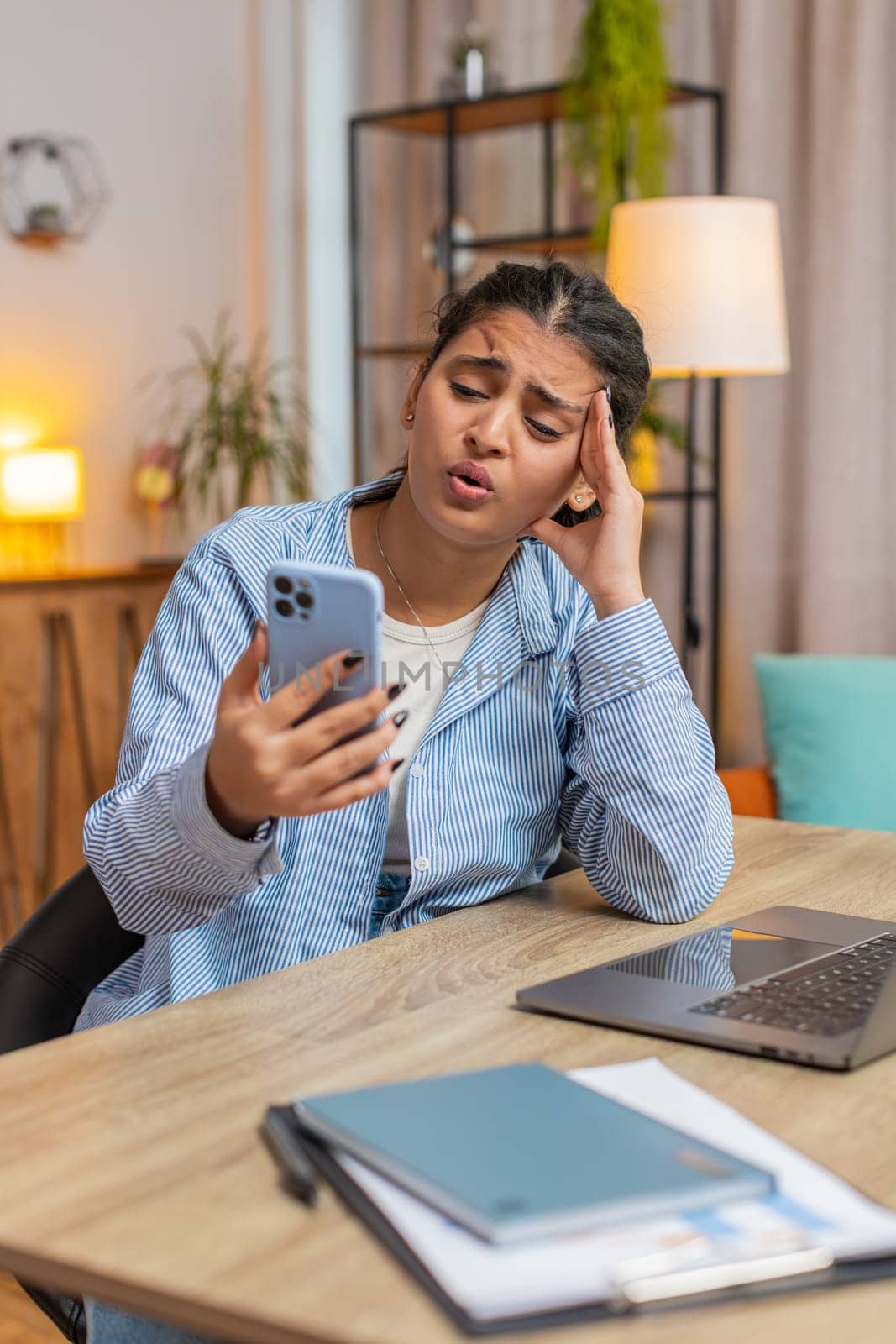 Upset displeased Indian woman freelancer using smartphone sitting at desk table in living room. Sad young girl touching head loses becoming surprised by sudden lottery results or bad news. Vertical.