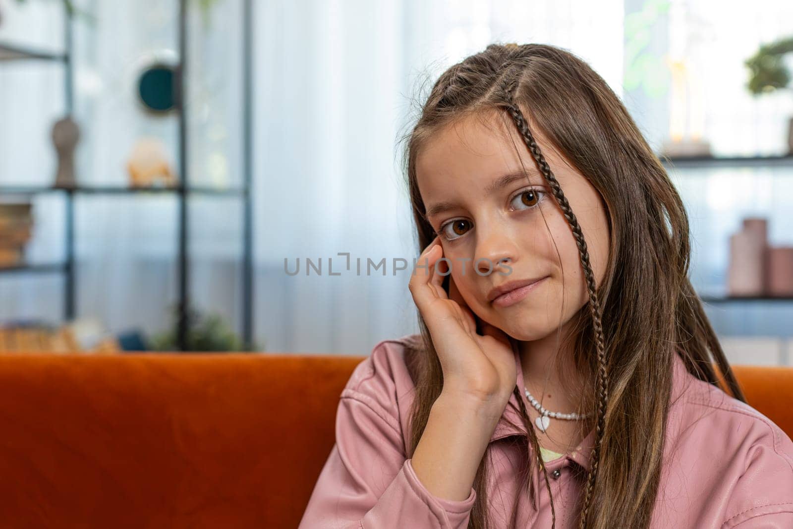 Portrait of happy smiling young girl child sitting on comfy sofa couch looking at camera in living room at home apartment. Cute teenage female school kid with brown long hair. Lifestyles.