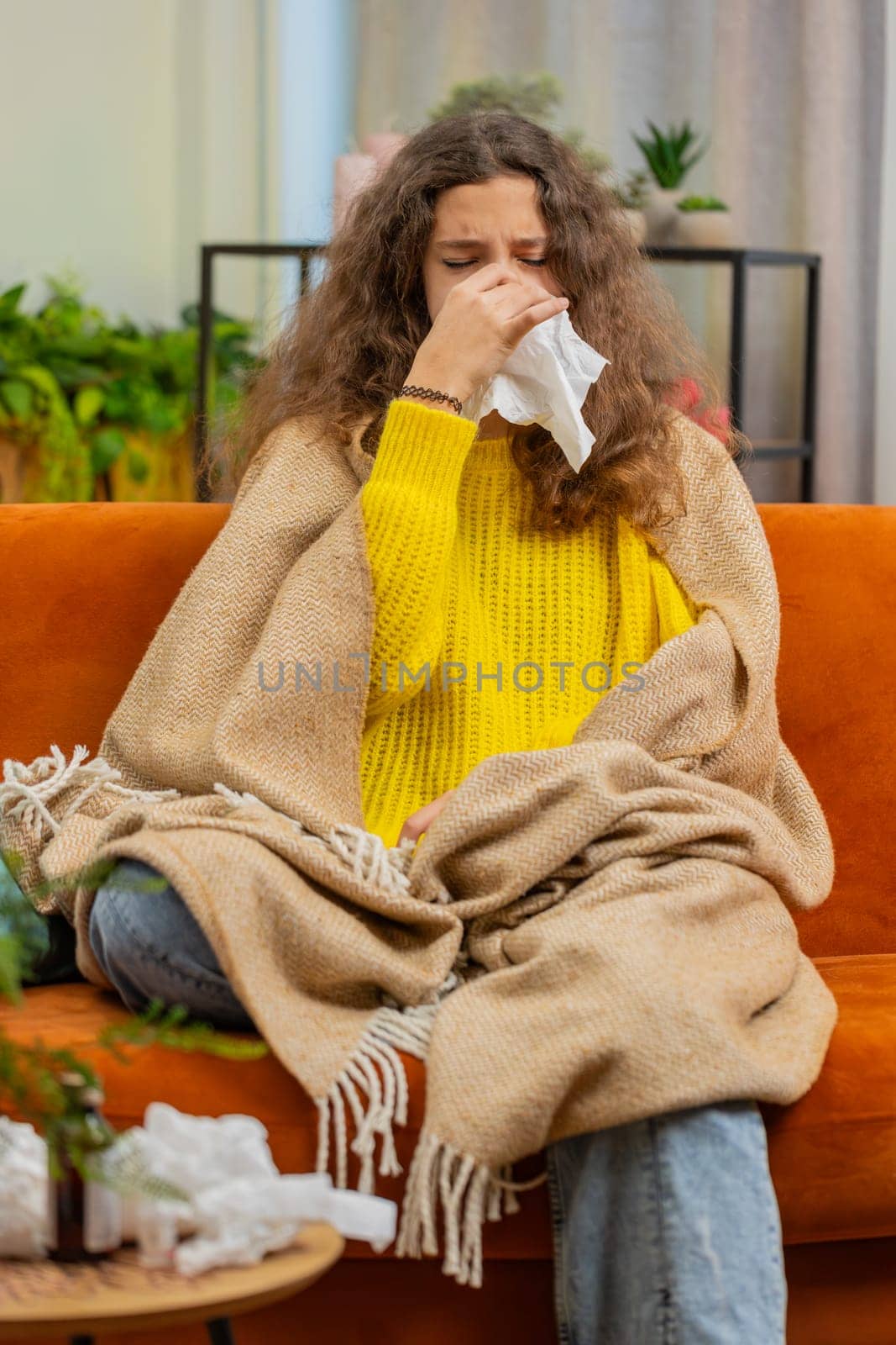 Ill child girl suffering from cold flu allergy infection sitting on sofa in living room at home. Sick teenager kid blows sneezes wipes snot into napkin. Coronavirus quarantine pandemic. Vertical