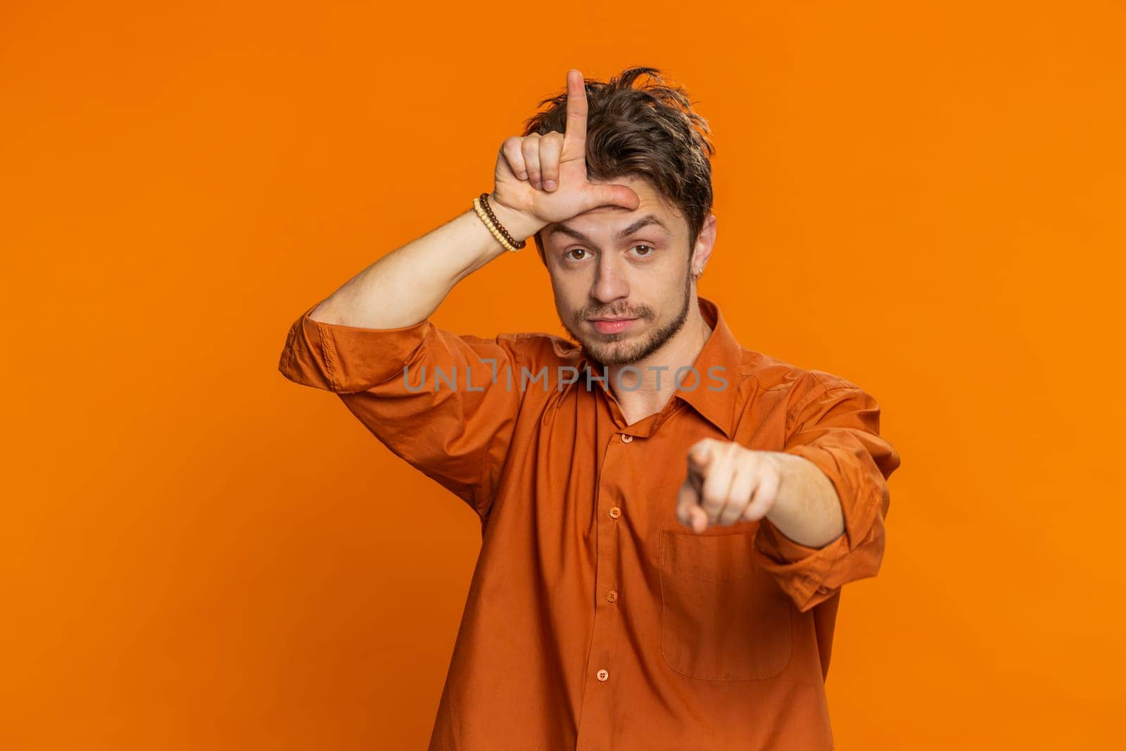 Caucasian man showing loser gesture and pointing on you, blaming accusing for unsuccess, expressing disrespect, mocking your failure, bullying abuse. Young guy isolated on orange background indoors