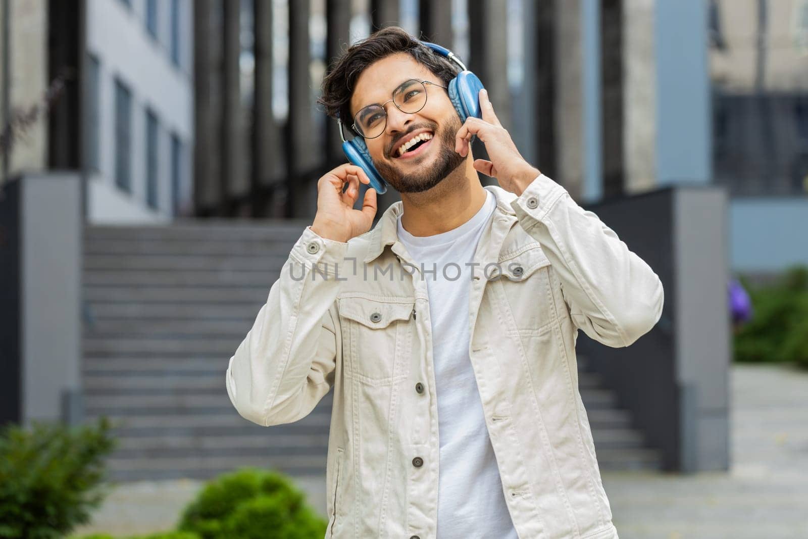 Happy relaxed young Indian man in wireless headphones dancing, listening favorite energetic disco music dancing outdoors. Arabian Hindu overjoyed guy standing in downtown city street. Business people
